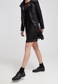 Schwarzes Spitzenkleid mit eng anliegendem Oberteil und ausgestellt Rock, kombiniert mit einer schwarzen Lederjacke und klobigen schwarzen Schnürschuhen.