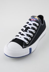 Svart canvas-sneaker med vita snören, vit gummikappa och blå detaljer. Har ögla och en logotyp-patch på tungan.