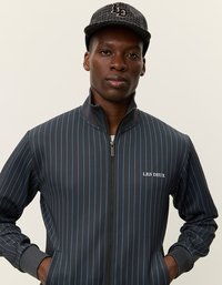 Chaqueta negra con cremallera y finas rayas verticales azules, con el logotipo de "LES DEUX". Se lleva con una gorra con estampado. Textura suave, diseño atlético.