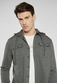 Szary, długi rękaw, koszula z kapturem, z dwiema kieszeniami z przodu i zapięciem na guziki. Noszona na gładkim białym t-shircie. Gładka tekstura.