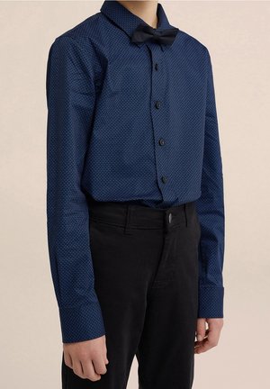 Enfant portant une chemise bleue à motifs à manches longues avec des boutons noirs et un nœud papillon noir, associée à un pantalon noir.