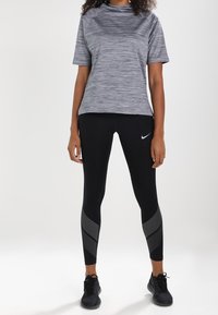 Top deportivo gris de manga corta con cuello alto, combinado con leggings negros que presentan un acento de rayas diagonales. Zapatillas de correr negras completan el conjunto.