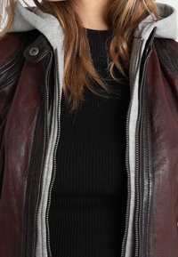 Braune Lederjacke mit einer grauen Kapuze, ausgestattet mit einem Reißverschluss und einem schwarzen, gerippten Sweater darunter. Auffällige Nahtdetails an den Schultern.