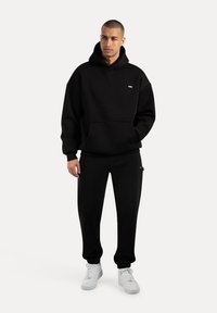 Mann trägt einen schwarzen Hoodie und passende schwarze Jogginghosen, steht mit den Händen in den Taschen und weißen Turnschuhen vor einem einfarbigen Hintergrund.
