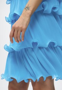 Κοντινό πλάνο χεριού με τατουάζ ήλιου και σελήνης να ακουμπά κοντά σε υφή ελαφρού μπλε υφάσματος με βολάν και πιέτες.