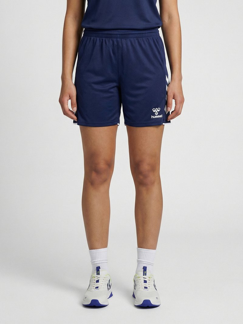 Person står iført marineblå træningsshorts med hvidt logo til venstre, hvide sokker og hvide sneakers med blå detaljer.