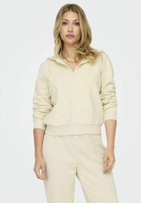 Felpa zip in beige chiaro realizzata in tessuto morbido, dotata di tasca a marsupio e polsini a coste. Abbinata a pantaloni coordinati in un taglio rilassato.