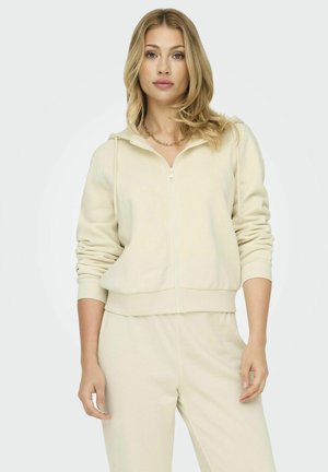 Felpa zip in beige chiaro realizzata in tessuto morbido, dotata di tasca a marsupio e polsini a coste. Abbinata a pantaloni coordinati in un taglio rilassato.