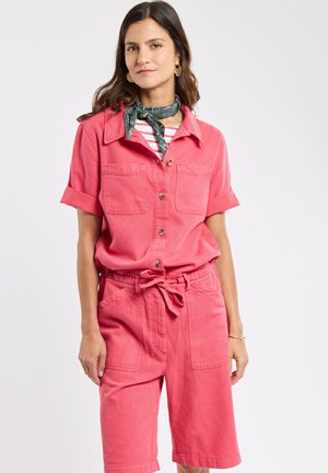 Femme portant une chemise rose à manches courtes boutonnée et un short assorti avec une taille nouée ainsi qu'un foulard vert autour du cou.