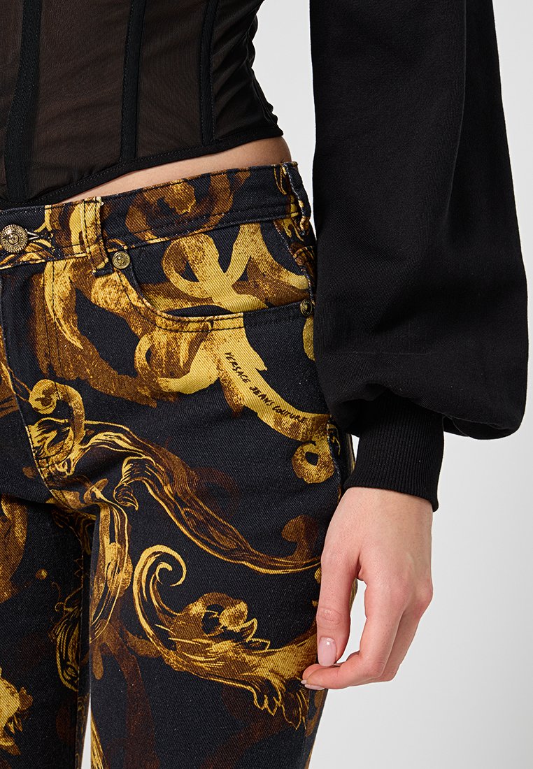 Top negro con mangas abullonadas combinado con jeans negros que presentan un patrón ornamentado en dorado; incluye dos botones metálicos en la cintura.