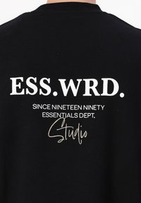 Black sweatshirt featuring white text: "ESS.WRD. SINCE NINETEEN NINETY ESSENTIALS DEPT." with a gold signature "Studio". Regular fit, soft texture.