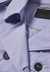 Gros plan d’un col de manteau violet clair boutonné montrant des boutons noirs et une étiquette noire à l’intérieur du col marquée "C&A".