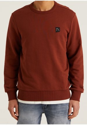 Hombre con sudadera de cuello redondo de color óxido con un pequeño parche negro con logo en el pecho, superpuesta sobre una camisa blanca y jeans azul claro.