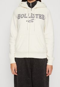 Krämfärgad zip-up-hoodie med "HOLLISTER"-logotyp i mörkblått. Har en fodrad huva, framfickor och ribbade muddar. Mjuk tygstruktur.