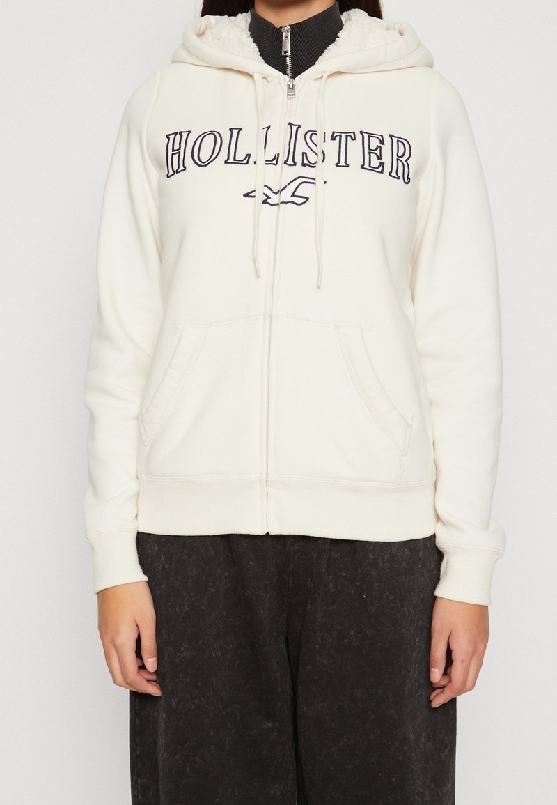 Krämfärgad zip-up-hoodie med "HOLLISTER"-logotyp i mörkblått. Har en fodrad huva, framfickor och ribbade muddar. Mjuk tygstruktur.