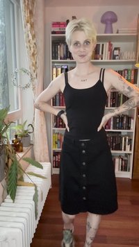 Pessoa com cabelo loiro curto e tatuagens usa uma blusa de alças preta e uma saia preta com botões, está de pé numa sala com estantes de livros e plantas.