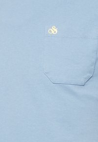 Vaaleansininen t-paita, jossa on tasku. Taskussa on pieni keltainen logo. Valmistettu pehmeästä puuvillamateriaalista, jolla on sileä pinta.