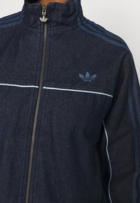 Mörkblå zip-jacka med ljusblå Adidas-logotyp på bröstet, tre ljusblå ränder på ärmarna och en metalldragkedja med logotyp.