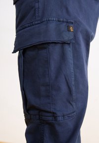 Granatowe spodnie cargo wykonane z bawełny, z bocznymi kieszeniami i wytrzymałym przeszyciem. Widoczna metka marki na kieszeni. Tekstura jest gładka i trwała.