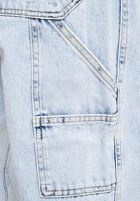 Lyseblå denimstoff med en litt falmet tekstur. Har fremtredende sømmer og en firkantet lommepatch med synlige sømdetaljer.