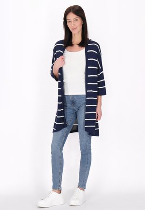 Femeie în picioare purtând un cardigan în dungi albastre și albe, top alb, blugi albaștri și adidași albi pe un fundal alb simplu.