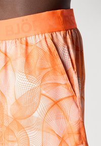 Oranžas peldbikses ar ģeometrisku acu rakstu, elastīgu jostu ar iegravētu "BJÖRN" tekstu un vieglu audumu.
