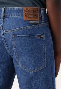 Blue denim jeans with a textured finish, featuring a brown leather back patch and stitched pocket details. Logo displayed on the fabric.
