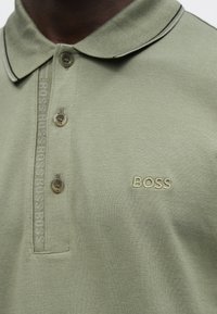 Zbliżenie jasnizeleńskiej koszulki polo z zapinanym na guziki kołnierzykiem i haftowanym na piersi napisem "BOSS".