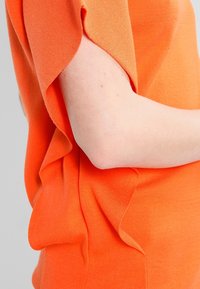 Pomarańczowy dzianinowy top o luźnym kroju z podwiniętymi rękawami i miękką fakturą, ukazujący delikatną tkaninę i subtelne detale drapowania.