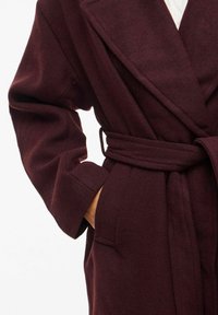 Burgundy wool wrap coat with large lapels, long sleeves, and a tied belt. Features side pockets and a soft texture.