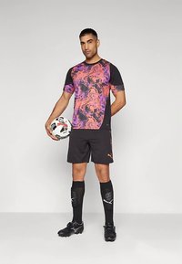 Mann iført svart og lilla mønstret fotballdrakt, holder en fotball, står med svarte sokker, leggbeskyttere og fotballsko på grå bakgrunn.