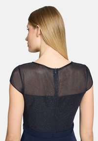 Robe bleu marine avec tissu transparent scintillant sur les épaules, col rond, et fermeture éclair discrète au dos. Texture douce et coupe ajustée.