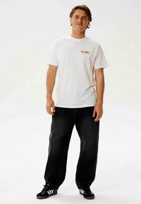 Camiseta blanca de algodón con un pequeño gráfico en naranja. Se combina con pantalones vaqueros negros de corte holgado y zapatillas deportivas negras con rayas.