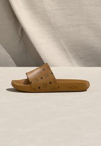 Sandalia tipo slide de cuero marrón con logos estampados en negro en la correa y logo MCM en relieve en el lateral, colocada sobre una superficie beige con un fondo de tela drapeada.