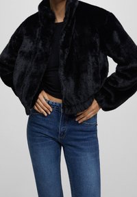 Kvinde iført en sort fuzzy zip-up jakke, sort top og blå denim jeans med hænderne hvilende på hoften mod en ensfarvet baggrund.