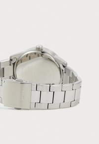 Reloj de pulsera de metal plateado con una pulsera de eslabones y un cierre grabado con el nombre de la marca "GUESS", mostrado desde la parte trasera.