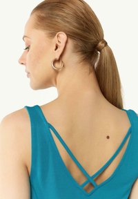 Turkusowy top sportowy z głębokim dekoltem w kształcie litery V i krzyżującymi się ramiączkami. Materiał ma gładką fakturę, a na skórze jest mała brązowa plama.