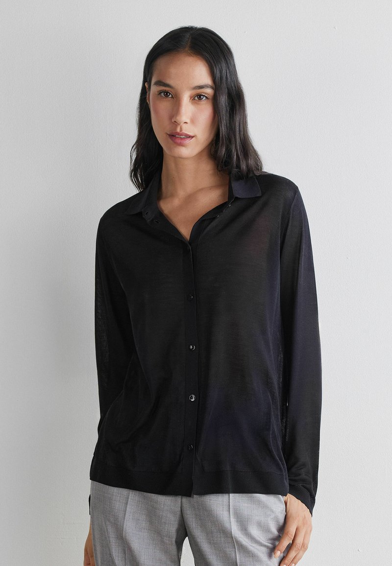 Vrouw met lang donker haar, gekleed in een zwarte doorgestikte cardigan en lichtgrijze broek, staande tegen een effen lichte muur.