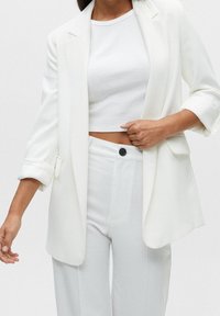 Blazer blanco ajustado con mangas enrolladas, combinado con un top blanco corto y pantalones de talle alto a juego. Tejido suave, diseño minimalista.