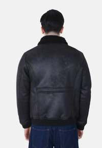 Uomo in giacca di shearling nera con polsini e orlo a coste, in piedi di spalle, abbinata a jeans di denim blu scuro.