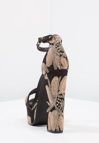 Sandalia negra de tacón alto con un patrón floral dorado texturizado, tira en el tobillo y una plataforma robusta para mayor altura.