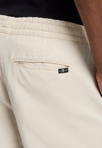 Beige shorts met een elastische tailleband en een achterzak. De stof lijkt glad met een subtiele glans. Inclusief een klein merkembleem.