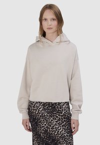 Vrouw draagt een beige cropped hoodie met rafelige randen en een zwarte en beige luipaardprint rok, staand tegen een effen achtergrond.