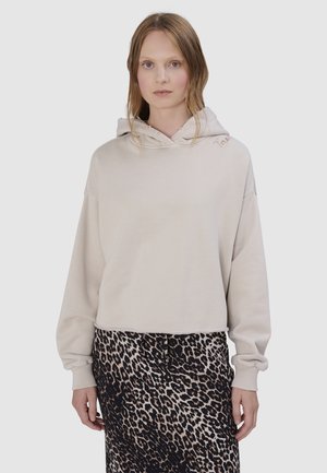 Vrouw draagt een beige cropped hoodie met rafelige randen en een zwarte en beige luipaardprint rok, staand tegen een effen achtergrond.