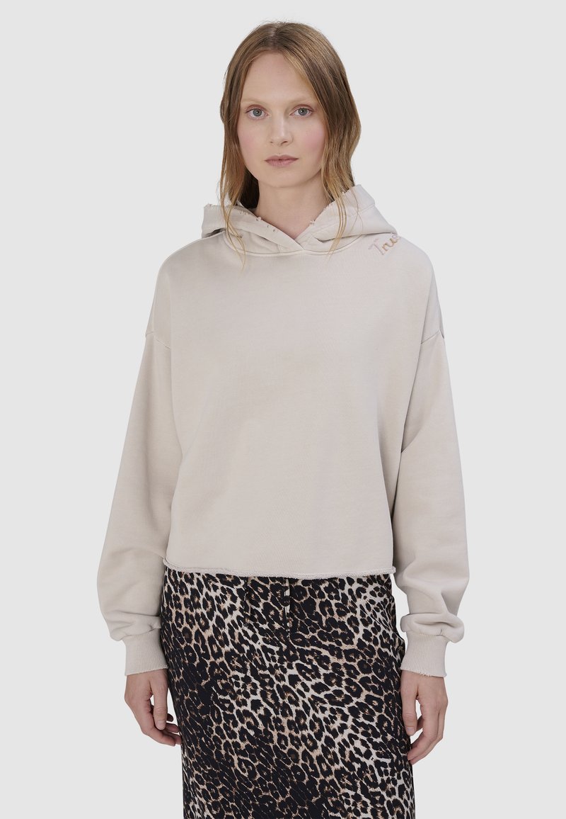 Vrouw draagt een beige cropped hoodie met rafelige randen en een zwarte en beige luipaardprint rok, staand tegen een effen achtergrond.