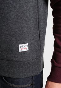 Sudadera gris hecha de una tela suave que presenta un dobladillo acanalado y una etiqueta de marca blanca con texto rojo en la parte inferior izquierda. Manga de color granate oscuro.