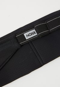 Musta juoksuvyö, jossa on teksturoitu kangas, heijastinauha ja HOKA-logo. Joustava hihna säädettävään istuvuuteen.