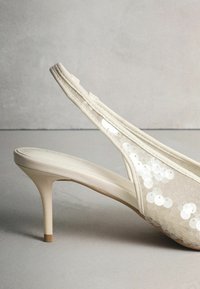 Smėlio spalvos aukštakulnis su atvira kulno dalimi, puoštas blizgučiais ant skaidrios medžiagos, su smailiomis noselėmis ir lieknu stiletto kulnu.