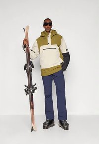 Skidklädsel med en beige och olivgrön jacka med en framficka, marinblå byxor, svarta skidpjäxor och skidor i handen.