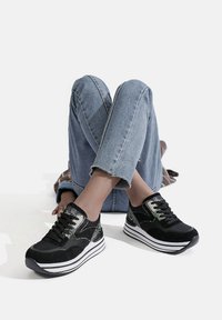 Sneaker neri e verdi con una tomaia in suede, design con lacci e suola bianca elevata con strisce nere. Indossati con jeans blu strappati.